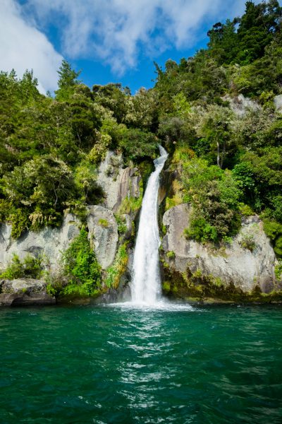 A waterfall in the bush of New Zealand, surrounded by lush greenery and blue sky, with water cascading down into turquoise waters below. The scene is captured from a boat on one side of the river looking towards the viewer. A large rock wall on the right forms part of the waterfall's drop point. Water splashes against rocks as it falls. There is some mist rising off the water near where its surface meets the air above the waterfall. Shot taken with a Canon EOS camera, wide angle lens, f/8 aperture setting, in the style of landscape photography. --ar 85:128