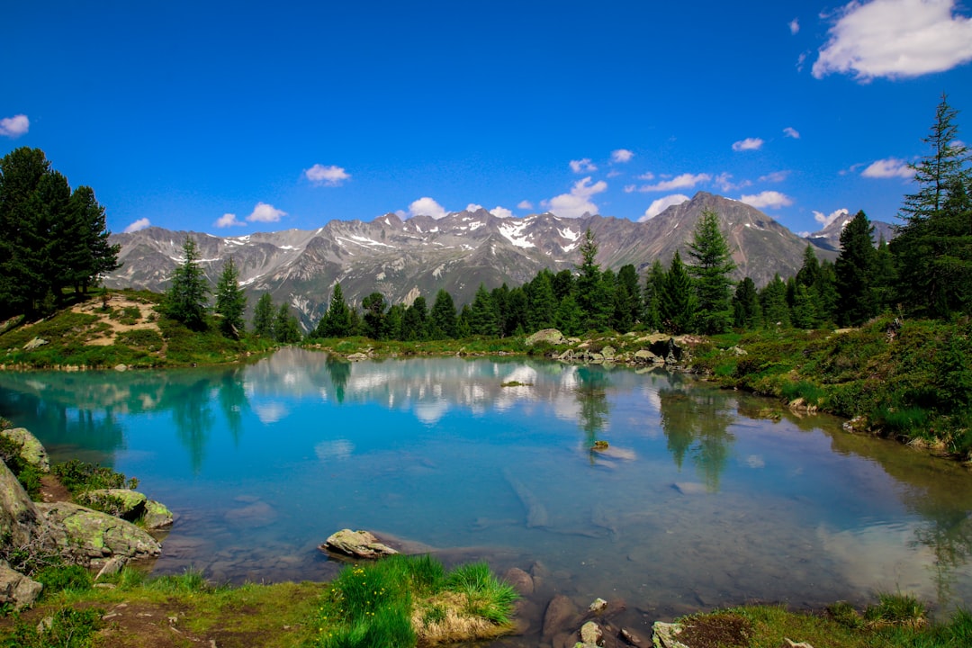 A lake in the Italian Alps with trees and mountains in the background, a clear blue sky, the water is a turquoise color, green grass on the ground, photo realistic in the style of canon eos r5. –ar 128:85