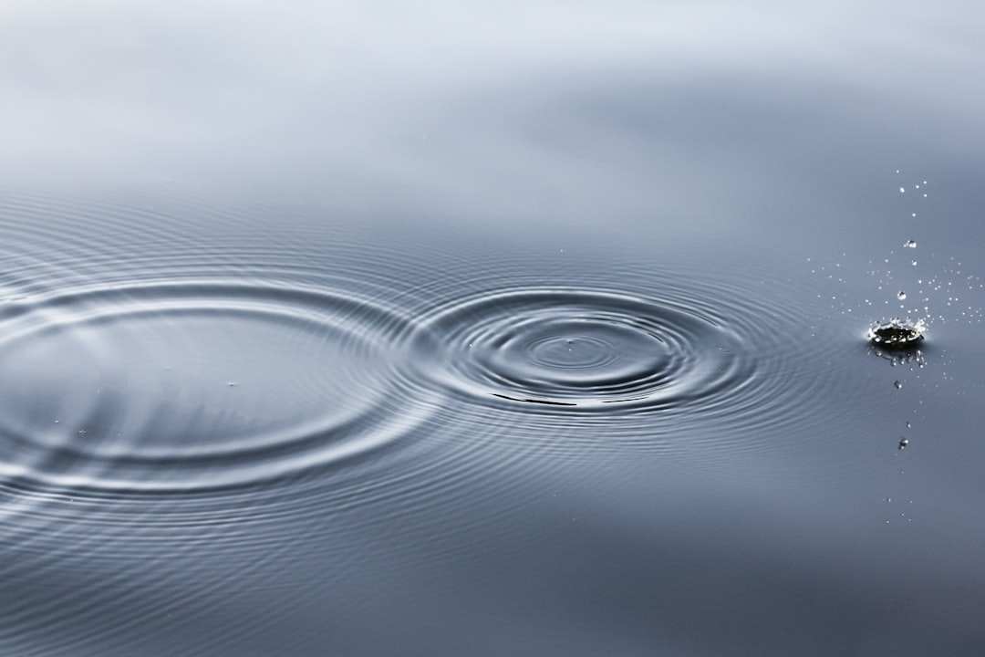 A photo of small ripples in the water, with one drop creating circular waves on the calm grey surface. The focus is sharp and clear, capturing the intricate details of each ripple’s shape and size. This scene evokes tranquility and harmony as these gentle splashes create delicate patterns across the still waters in the style of calm waters. –ar 128:85