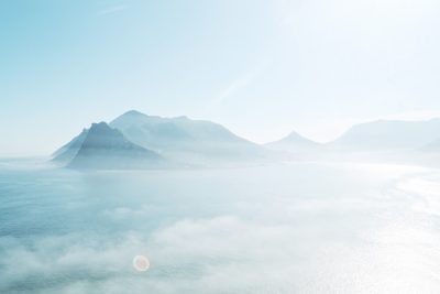 Beautiful foggy view of Cape Town's island, mountains in the distance, sun shining through clouds, white sky, light blue sea, shot from high above, minimalist photography, high definition, high resolution, cinematic, high detail. --ar 128:85