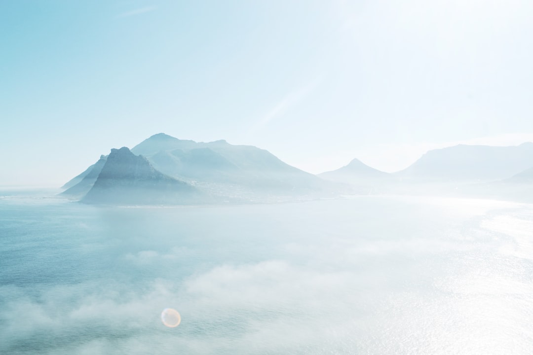 Beautiful foggy view of Cape Town’s island, mountains in the distance, sun shining through clouds, white sky, light blue sea, shot from high above, minimalist photography, high definition, high resolution, cinematic, high detail. –ar 128:85