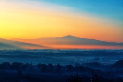 photograph of the sunrise over an italian landscape with mountains in the background, misty morning light, blue and orange sky, distant hills, distant trees, distant buildings, distant village, distant mountain range, distant peak, shot on canon eos r5 --ar 128:85