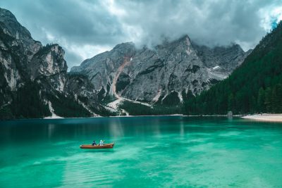 photograph of Lake Braies in the Dolomites with turquoise water and mountains, people on a rowing boat in the background, cinematic, cloudy sky, green colors, photography in the style of unsplash, shot from a drone, --ar 128:85