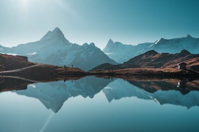 A photo of the Swiss Alps with mountains reflecting in a lake, clear sky, daytime, blue tones, people hiking around the mountain. The photo is in the style of a landscape painting. --ar 128:85