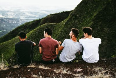 Four young men sitting on the edge of an alpine cliff, overlooking green hills in Hawaii. One man is holding his arm around another's shoulder for support and comfort as they look out over their surroundings together. Shot from behind them with a wideangle lens to capture both of each person and convey friendship among all four friends. Soft natural lighting highlights facial expressions of joy against the backdrop of nature's grandeur. --ar 128:85