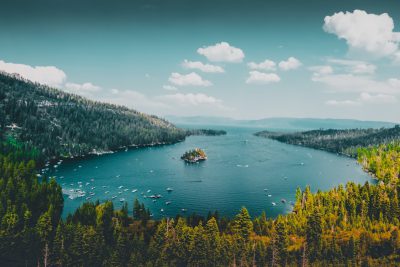 panoramic view of lake tahoe, lake emerald in the distance with small island and boats, forest on mountainsides around it, blue sky with clouds, unsplash photography style, --ar 128:85