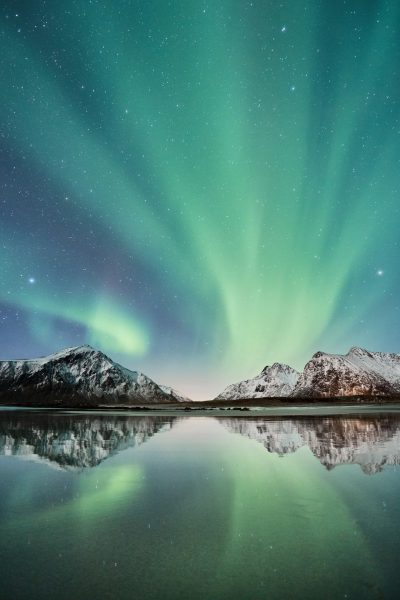 A beautiful photo of the aurora borealis, with snow-covered mountains reflecting in water and stars shining in the sky. The green light from the polar lights is reflected on the surface of the lake, creating a stunning effect. Nikon D850 camera, f/2 lens, 36mm focal length, aperture at f/4 for sharpness. Low angle shot to capture the grandeur of the scene. --ar 85:128