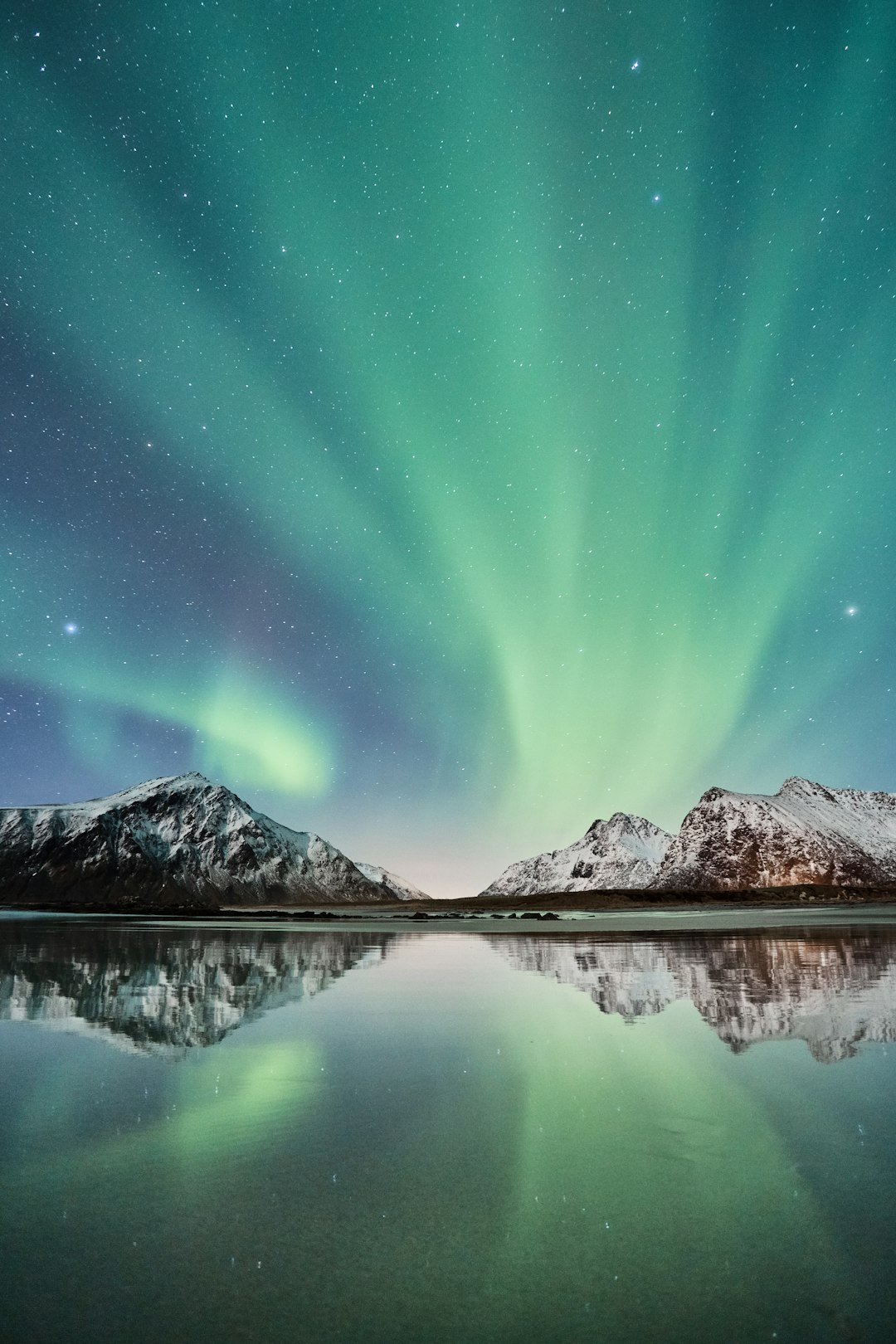 A beautiful photo of the aurora borealis, with snow-covered mountains reflecting in water and stars shining in the sky. The green light from the polar lights is reflected on the surface of the lake, creating a stunning effect. Nikon D850 camera, f/2 lens, 36mm focal length, aperture at f/4 for sharpness. Low angle shot to capture the grandeur of the scene. –ar 85:128