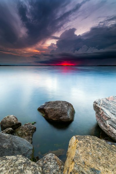 A serene lakeside at sunset, with rocks and reflections in the water, under dark stormy clouds. The sky is painted with hues of red and purple as the sun sets behind the horizon, casting long shadows over calm waters. In the style of sharp focus photography. --ar 85:128