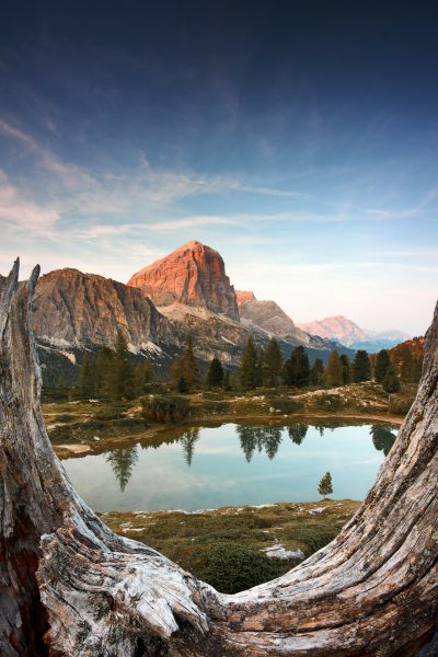 A mountain lake in the Dolomites with an impressive mountain peak in the background, seen through tree trunk frames, summer time, blue sky, sunset light, shot in the style of a National Geographic magazine photographer. --ar 85:128