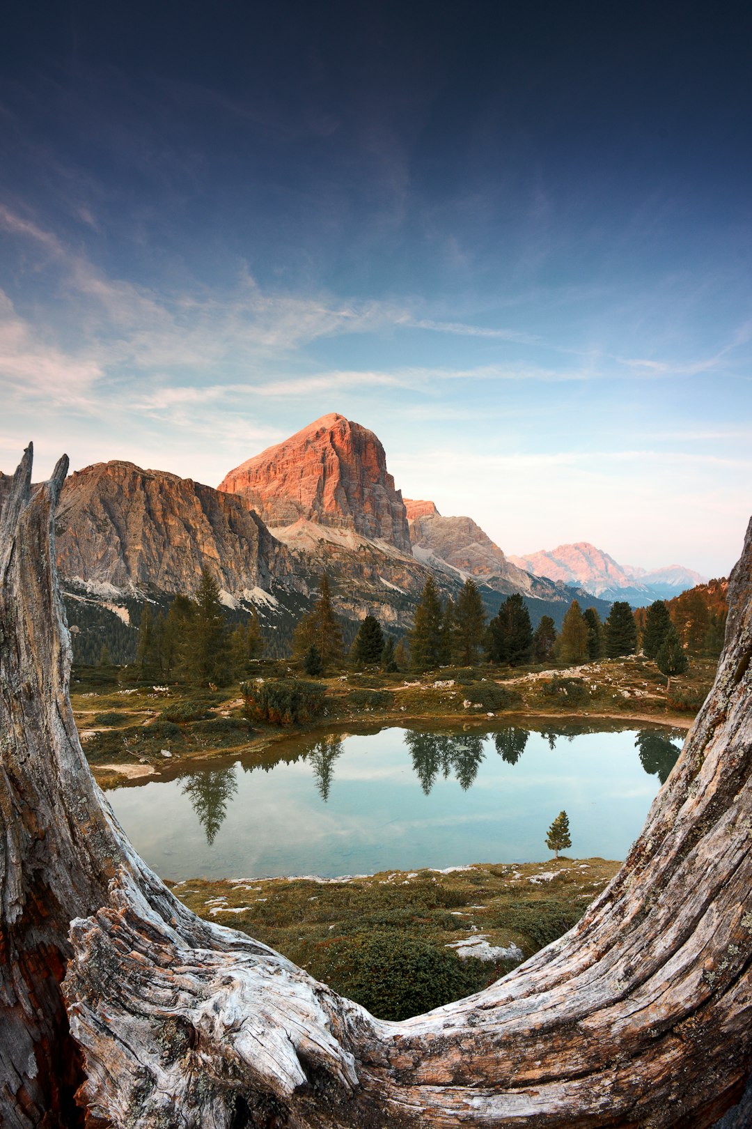 A mountain lake in the Dolomites with an impressive mountain peak in the background, seen through tree trunk frames, summer time, blue sky, sunset light, shot in the style of a National Geographic magazine photographer. –ar 85:128