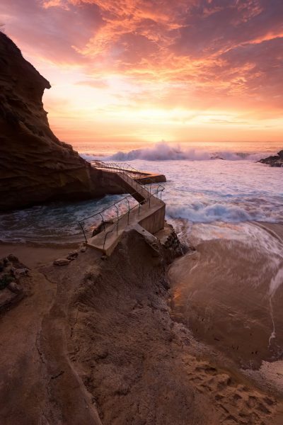 Photograph of the natural pool at La Jolla, California with a beach and waves during sunset. The water is calm and there's an old wooden bridge leading to it from above. There's a pink sky with orange clouds in the background. In the foreground you can see sand on one side and rocks on another. A small path leads up to where stairs lead into the ocean. This photo was taken using a Canon EOS R5 camera with a wideangle lens in the style of a landscape photographer. --ar 85:128