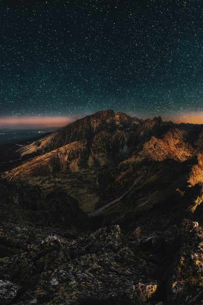 Cinematic photo of the Tatra Mountains at night, with stars in the sky and a glow from an iPhone on top of one mountain. The scene is illuminated by moonlight, creating deep shadows between rocks and mountains, with subtle light effects adding depth to the composition. In closeup view, the rugged landscape appears mystical under starry skies in the style of moonlight. --ar 85:128