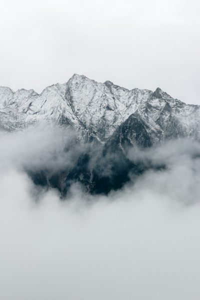 Snowy mountains surrounded by clouds, a distant view of snowcapped peaks shrouded in mist and fog. A minimalist photography style with a white background, simple composition, high definition photography, clean and clear focus, detail rendering, low saturation, large aperture, soft light. The black mountain is located at the bottom left corner of the screen, with only half visible above it covered in heavy white cloud and mist. It's a beautiful scenery. High resolution, high quality. --ar 85:128