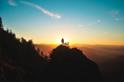 A silhouette of an adventurer standing on top of the mountain, overlooking beautiful scenery at sunrise. The sky is clear and blue with some clouds. In front there is another high hill covered in trees. There is also one small tree growing from between two rocks on that slope. On its branches stands a person holding out their hand to touch something above them, in the style of Canon EOS R5 camera with wideangle lens. --ar 128:85