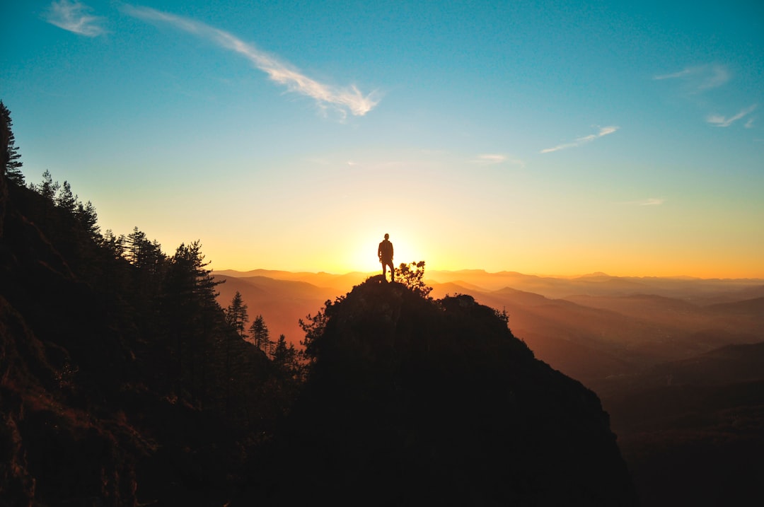 A silhouette of an adventurer standing on top of the mountain, overlooking beautiful scenery at sunrise. The sky is clear and blue with some clouds. In front there is another high hill covered in trees. There is also one small tree growing from between two rocks on that slope. On its branches stands a person holding out their hand to touch something above them, in the style of Canon EOS R5 camera with wideangle lens. –ar 128:85