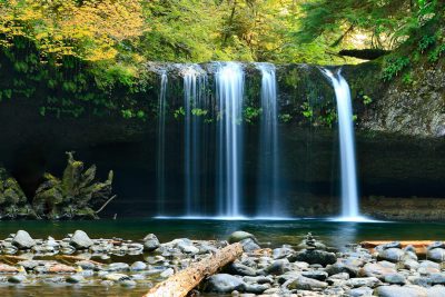 Photo of Silver Falls in Oregon, with water flowing over rocks and pebbles with lush greenery on the sides. A waterfall with two white vertical drops falling from top to bottom. A fallen log near the water's edge in a peaceful natural scene with natural light. --ar 128:85