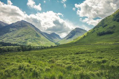 A green grassy valley with Scottish mountains in the background, a sunny day with a blue sky and clouds. Photographed with a Canon EOS R5 in the style of no particular artist. --ar 128:85