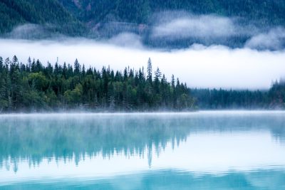 Photograph the misty beauty of Lakeplei in British Columbia, Canada with its turquoise waters reflecting the surrounding pine forest and mountains. The fog adds an ethereal touch to create a serene scene perfect for adding a sense of tranquility or mystery to your designs. , shot by Sony Alpha A7 IV camera. ISOs from 200450, --ar 128:85