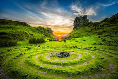 Beautiful landscape photo of the circular labyrinth in green grass with small rocks leading to an ancient Celtic stone cairn and bonfire at sunset, on top of a lush valley surrounded by rolling hills in a British island park with blue sky and white clouds, in the style of National Geographic photography, with a wide angle, epic, cinematic view with vibrant colors, following the rule of thirds composition during the golden hour sunset. --ar 128:85