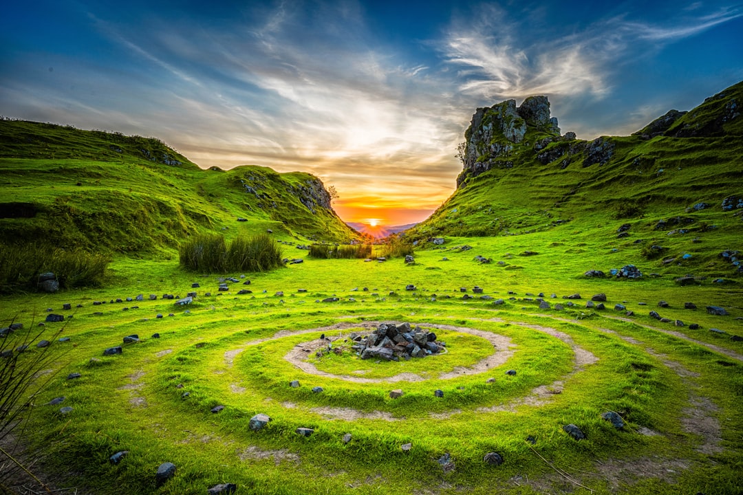 Beautiful landscape photo of the circular labyrinth in green grass with small rocks leading to an ancient Celtic stone cairn and bonfire at sunset, on top of a lush valley surrounded by rolling hills in a British island park with blue sky and white clouds, in the style of National Geographic photography, with a wide angle, epic, cinematic view with vibrant colors, following the rule of thirds composition during the golden hour sunset. –ar 128:85