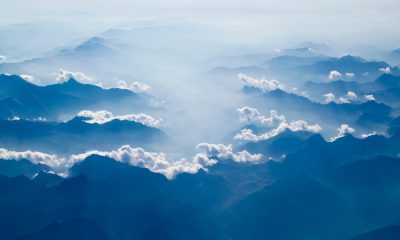 The aerial view of the mountains in China, with white clouds floating above and sunlight shining through on top of blue mountain peaks. The entire scene is captured from an airplane window perspective, creating a breathtaking natural landscape. This photograph was taken using a Canon EOS R5 mirrorless camera and Fujifilm Provia film to capture the vibrant colors and textures of these majestic terrain features, in the style of Chinese landscape paintings. --ar 32:19