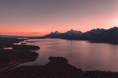 A drone photo of Grand Teton National Park at sunset, the lake is still and calm with beautiful pink hues in the sky. Mountains are on both sides of the water, trees cover half of it. The view from above captures the vastness of nature. Realistic photography shot in the style of National Geographic, taken with a Sony A7s iii camera and 35mm f/2 lens. --ar 128:85