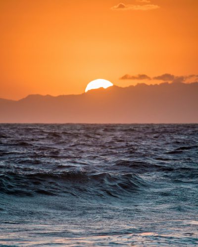 A sunset over the ocean with mountains in the background, orange sky, sun setting behind waves, horizon line, Hawaii island in the distance, photographed with a Sony A7 III camera using a 24-35mm f/8 lens. --ar 51:64