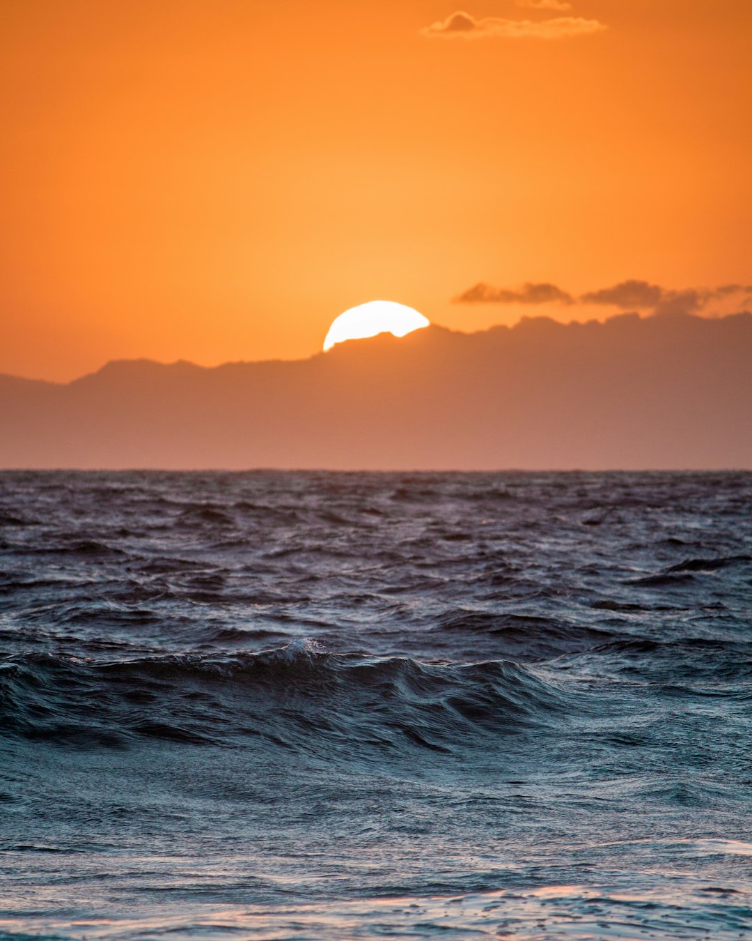 A sunset over the ocean with mountains in the background, orange sky, sun setting behind waves, horizon line, Hawaii island in the distance, photographed with a Sony A7 III camera using a 24-35mm f/8 lens. –ar 51:64