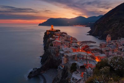 Vernazza, Cinque Terre at sunset with the village lights glowing in warm hues against the backdrop of rugged cliffs and an endless sea. The photograph captures the tranquil beauty as evening falls over the picturesque Italian coastline. Nikon D850 DSLR photo in the style of Nikon. --ar 128:85