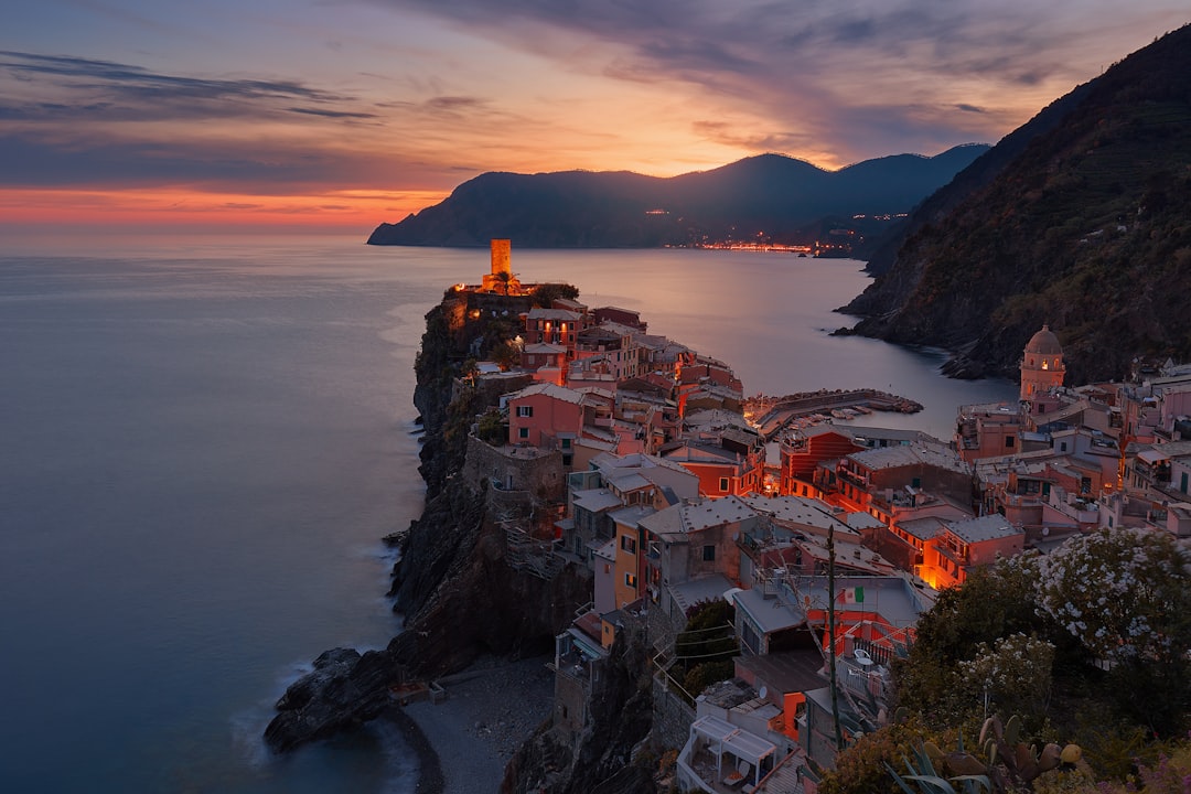 Vernazza, Cinque Terre at sunset with the village lights glowing in warm hues against the backdrop of rugged cliffs and an endless sea. The photograph captures the tranquil beauty as evening falls over the picturesque Italian coastline. Nikon D850 DSLR photo in the style of Nikon. –ar 128:85