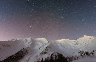 Starry sky over snowy mountains, a breathtaking view of the night sky filled with stars above snow-covered peaks in a winter mountain landscape. The vast expanse stretches as far up to celestial bodies like Jupiter and Saturn, creating an awe-inspiring scene that captures nature's beauty at its most serene. Captured in the style of a professional photographer using high-end camera equipment, the photograph showcases intricate details of star patterns against the backdrop of pristine white scenery. --ar 64:41