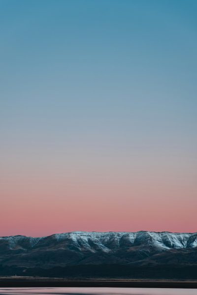 In the evening, there is snow on top of distant mountains under a clear sky with pink and blue tones. The foreground features water in front of it, and the sky takes up most space. It has a minimalist style and a high definition photography effect. --ar 85:128