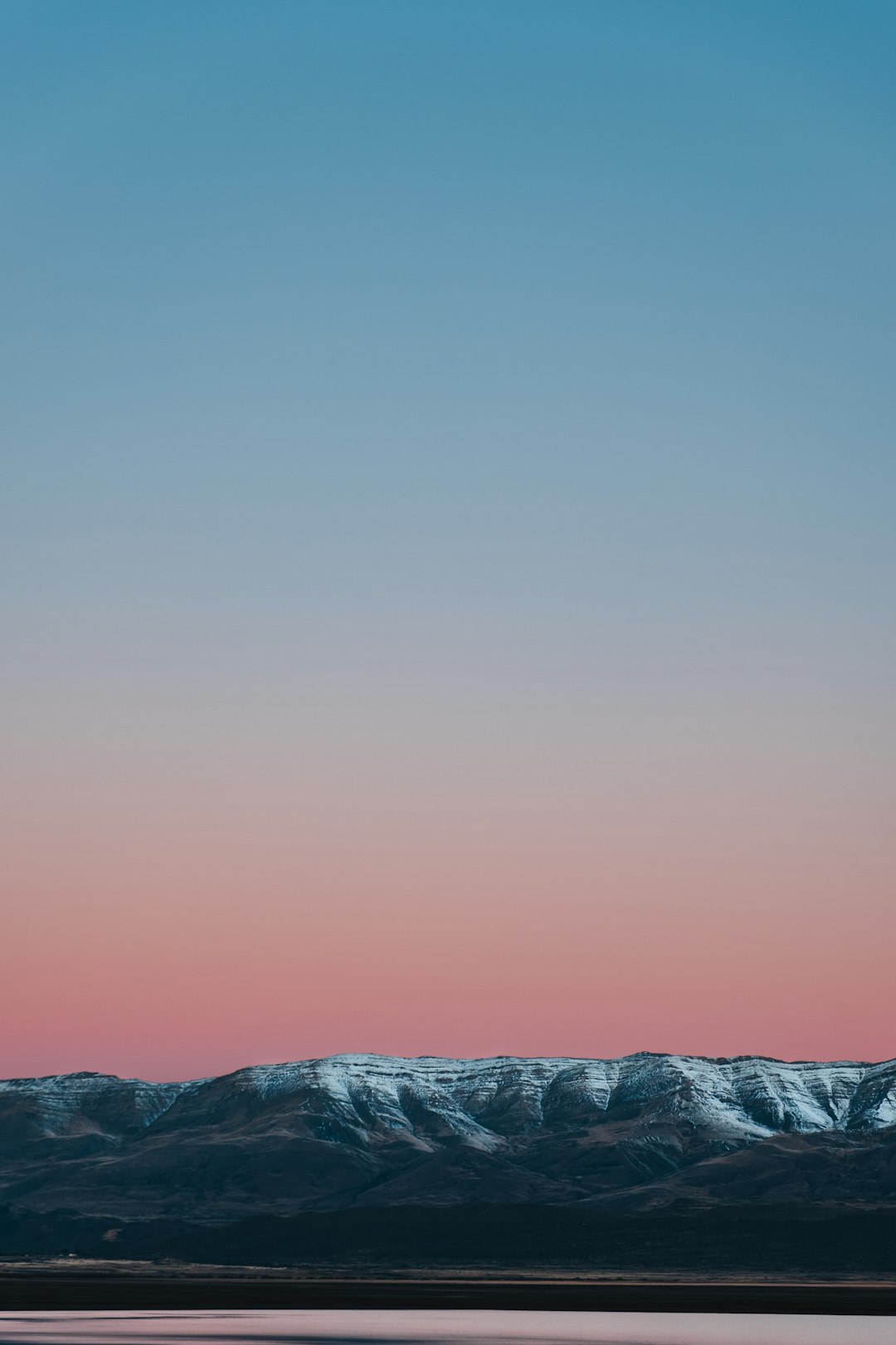 In the evening, there is snow on top of distant mountains under a clear sky with pink and blue tones. The foreground features water in front of it, and the sky takes up most space. It has a minimalist style and a high definition photography effect. –ar 85:128