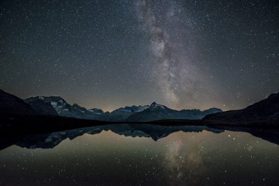 A starry night sky over the Swiss Alps, with the reflection perfectly in still water of Lake Blurry reflection. The Milky Way is clearly visible in the sky and there are no other stars that can be seen. It was taken from an eerie spot where you could only see the lake and mountains, so it feels like being alone at dawn. There were a few clouds but they all reflected on the lake, creating a beautiful contrast between them and the darkening sky. --ar 128:85