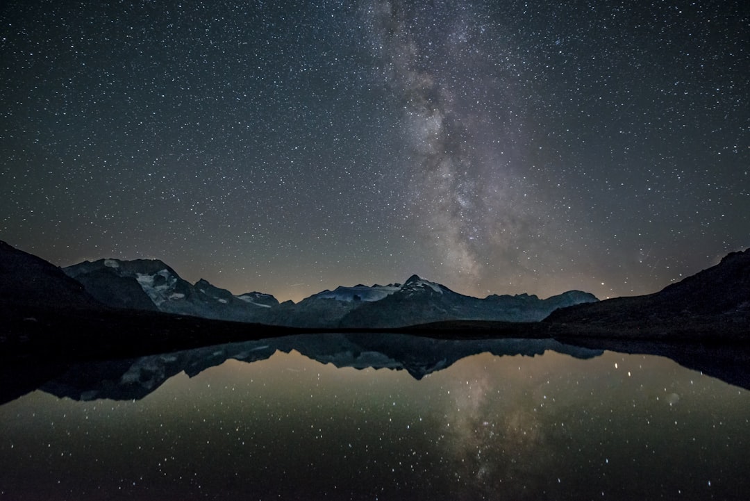 A starry night sky over the Swiss Alps, with the reflection perfectly in still water of Lake Blurry reflection. The Milky Way is clearly visible in the sky and there are no other stars that can be seen. It was taken from an eerie spot where you could only see the lake and mountains, so it feels like being alone at dawn. There were a few clouds but they all reflected on the lake, creating a beautiful contrast between them and the darkening sky. –ar 128:85