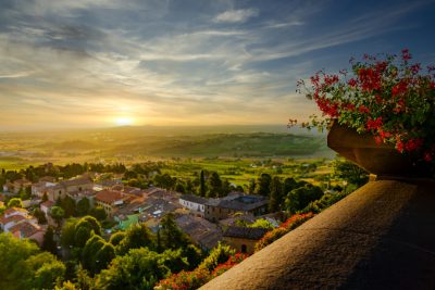 Sunrise view from Sang tadates tower overlooking the small town of Sog physically in Tuscany Italy, flower boxes on balcony with red flowers overlooking green rolling hills and distant white buildings, sun is rising over horizon casting long shadows, golden light illuminating sky, tranquil peaceful scene, warm sunny day, high resolution photography --ar 128:85