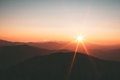 A beautiful sunrise over the mountains, with lens flare and a star in front. The sun is setting behind the silhouette mountain range. In front there's a forested valley, captured from an aerial perspective. A photo taken in the style of Sony Alpha a7 III camera using a wideangle lens at f/8 aperture setting. The colors of dawn create a warm atmosphere, and the sky conveys tranquility as sunlight illuminates the landscape. --ar 128:85