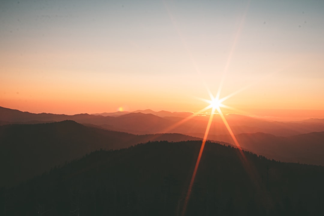 A beautiful sunrise over the mountains, with lens flare and a star in front. The sun is setting behind the silhouette mountain range. In front there’s a forested valley, captured from an aerial perspective. A photo taken in the style of Sony Alpha a7 III camera using a wideangle lens at f/8 aperture setting. The colors of dawn create a warm atmosphere, and the sky conveys tranquility as sunlight illuminates the landscape. –ar 128:85