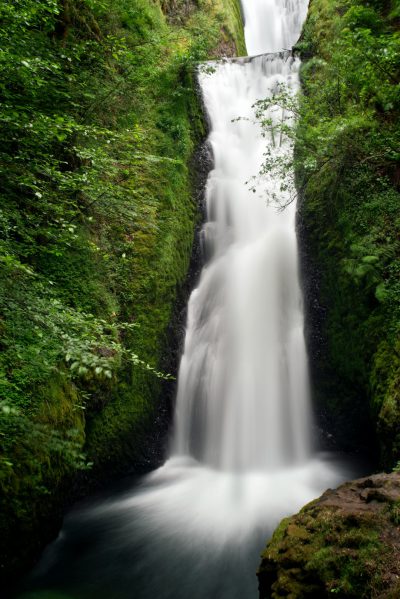 Photo of falling water in Hanes Gorge, Oregon, waterfall in the background, lush greenery, mossy rocks, long exposure, soft focus, natural light, peaceful atmosphere, wide angle lens, DSLR camera. --ar 85:128