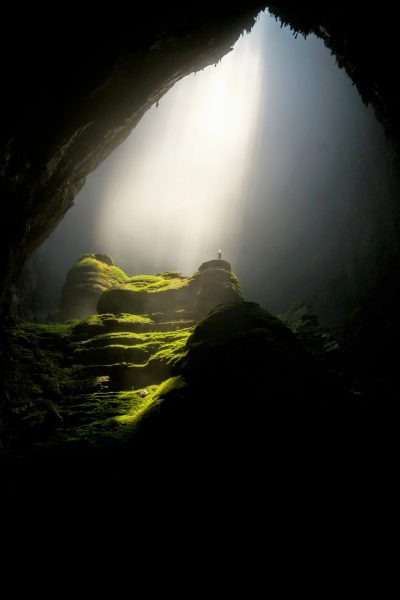 A photography of the inside view of a huge cave, with mossy rocks and green grass on top. Mist in the air, with sunlight shining through the sky from above. A tiny person standing far away in the dark mood. --ar 85:128