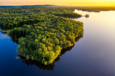 Aerial view of the Great Bass Perry forest island in Almanh Ngha Lake, New Jersey at sunset. The lake is surrounded by dense green forests and blue water with an isolated jungle-covered terrain island in the center. There is golden sunlight casting long shadows over the tranquil waters. Shot on Canon EOS R5 camera with RF60mm F2 wide angle lens. --ar 128:85