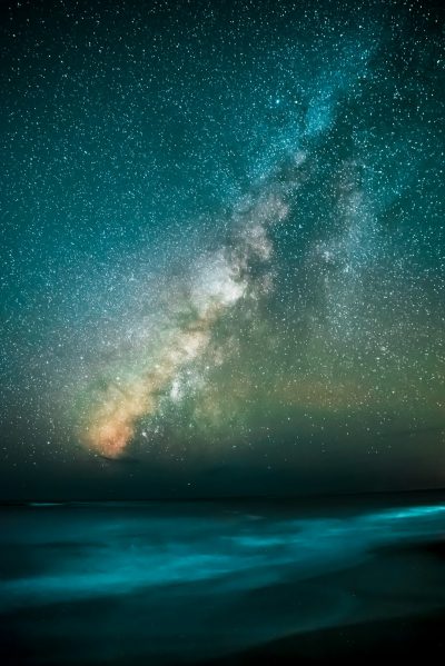 A long exposure photo of the Milky Way over an ocean in a starry night sky, with light painting in a dark teal and black color scheme. --ar 85:128