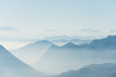 The light blue sky is surrounded by the misty mountains, creating an ethereal and dreamlike atmosphere. The distant peaks blend into each other in soft shades of gray against the backdrop of a clear sky. Aerial photography techniques capture the vastness of these mountainous landscapes, adding depth to the scene. This photo was taken with Canon EOS R5 using a wideangle lens at f/4 aperture setting, capturing every detail of the majestic landscape. --ar 128:85