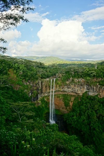 A wide shot of the waterfall in plateau, surrounded by lush greenery and dense vegetation. The sky is clear with fluffy white clouds adding to its picturesque beauty. In front stands Chamental Waterfall on its high cliff overlooking valley below. A panoramic view capturing the majestic height of chevents waterfalls against the backdrop of tropical landscape in the style of tropical landscape. --ar 85:128