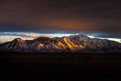 photo of the mountain range near Salt Lake City, golden hour lighting, dramatic sky, dark clouds on top with light coming from behind the mountains, snow covered peaks, shot in the style of Sony Alpha A7 III --ar 128:85