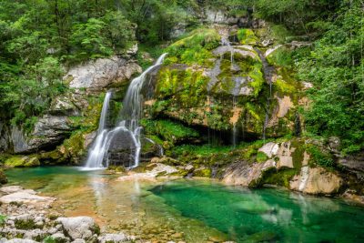 Waterfall in nature, forest and green rocks with water spring pool in the middle of waterfall. Travel background. Europe summer landscape. Velsor national park, Trigalav mountains area, Gaberrow am bobin potz under a clear sky. A view from behind the waterfall to the valley below, capturing its beauty. The scene is set against lush greens, adding depth and vibrancy to the overall composition. It's an ideal representation for travel printing or stock photography in the style of Nighton. --ar 128:85