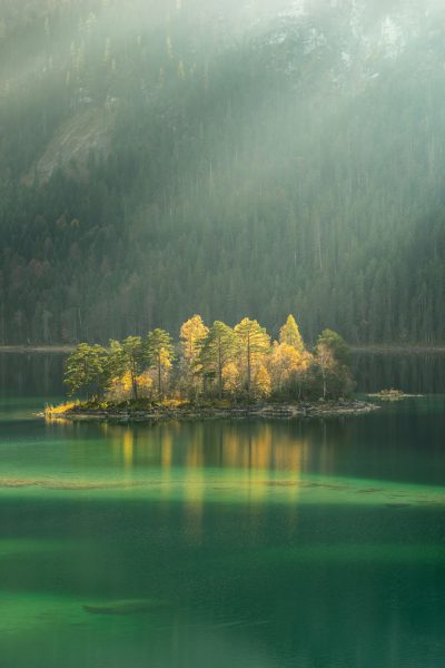 An island in the middle of Eibsee lake, light green water and yellow trees on it, sunlight through the foggy forest, landscape photography, canon eos r5 in the style of canon eos r5. --ar 85:128