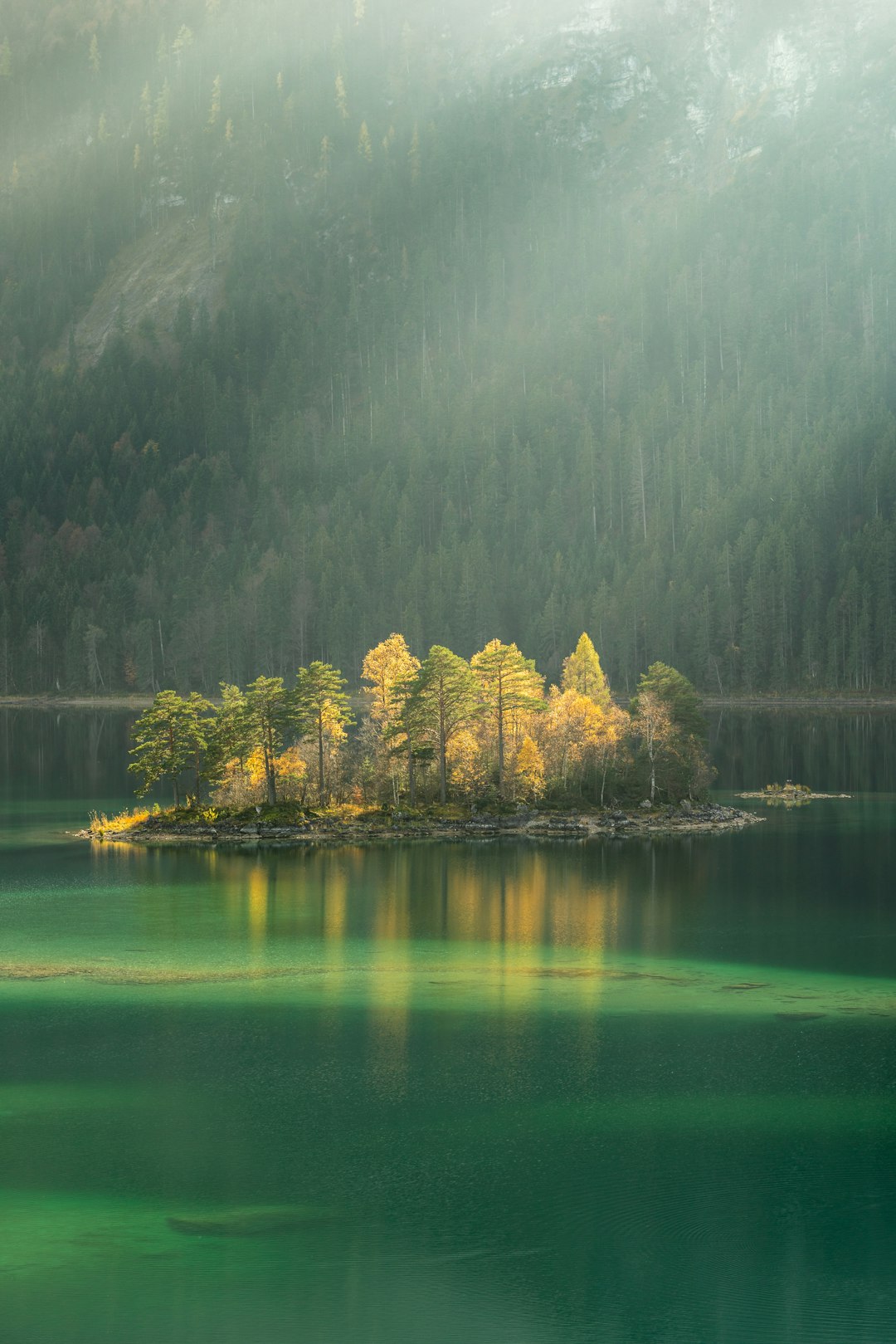 An island in the middle of Eibsee lake, light green water and yellow trees on it, sunlight through the foggy forest, landscape photography, canon eos r5 in the style of canon eos r5. –ar 85:128