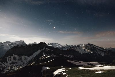 A panoramic view of the Swiss Alps at night, with stars and moonlight illuminating snowcapped peaks in an epic landscape. The scene is captured from high above in the style of camera raw. Shot on Fujifilm Pro 400H film stock for cinematic realism. --ar 128:85